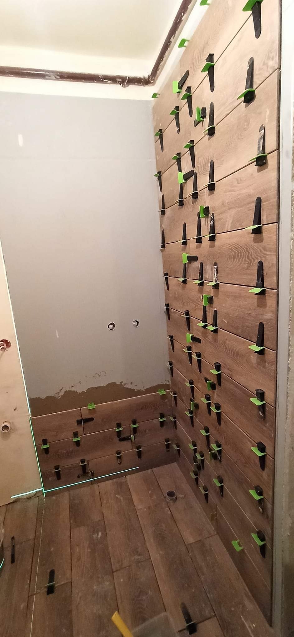 Montaż płytek imitujących drewno w łazience, z użyciem poziomicy laserowej i klipsów poziomujących, widoczne otwory na rury.