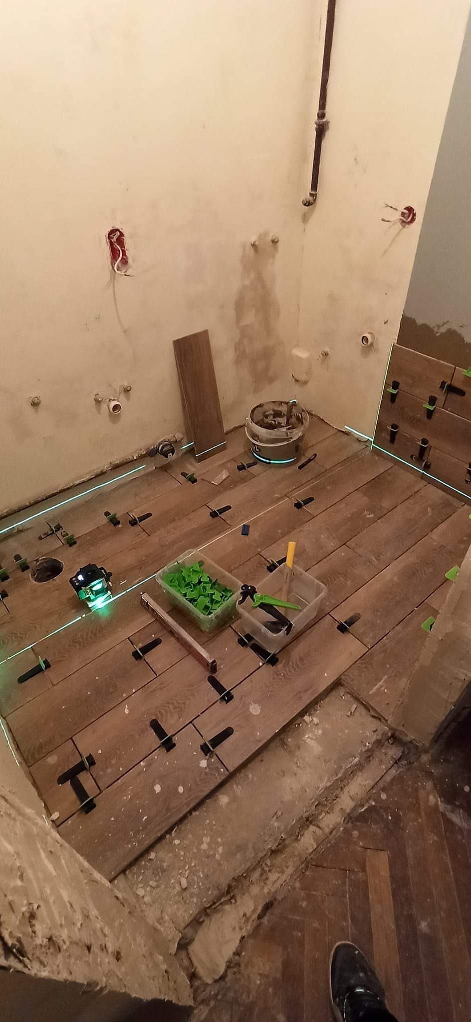 Podłoga z drewnopodobnych płytek w trakcie układania, z użyciem poziomicy laserowej i systemu poziomowania, widoczne akcesoria glazurnicze i surowe ściany z instalacjami.