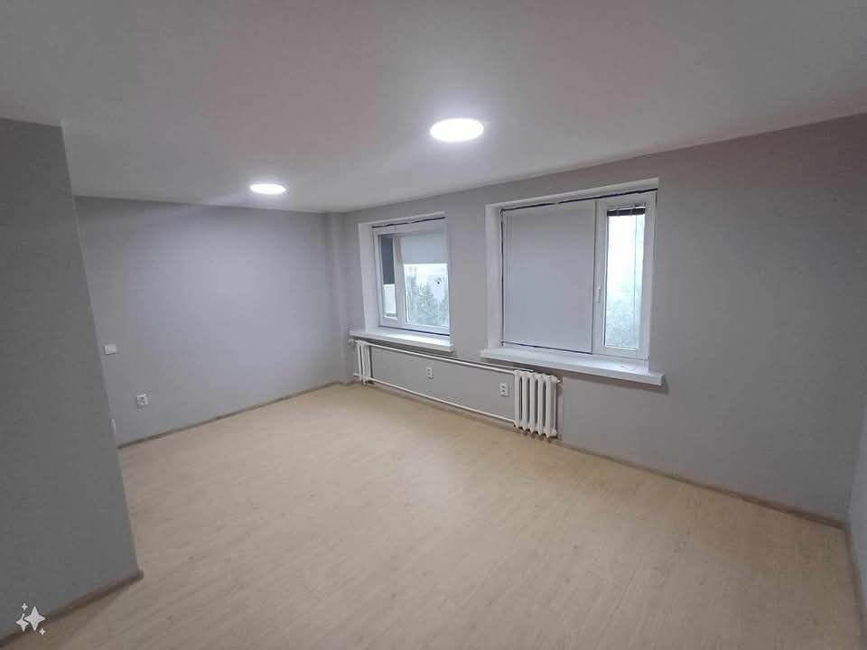 Pomalowane na szaro puste pomieszczenie z dwoma oknami z białymi ramami, jasnymi panelami podłogowymi i białymi grzejnikami pod oknami.