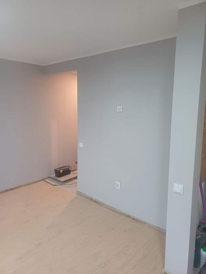 Jasnoszary pokój po malowaniu w Aleksandrowie Łódzkim, z drewnianą podłogą i skrzynką narzędziową w tle, widoczne gniazdka elektryczne i włączniki.