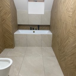 CER-MUR Przemysław Rosik - Nowoczesna łazienka na poddaszu z drewnopodobnymi płytkami na ścianach ułożonymi we wzór jodełki, beżowe płytki podłogowe imitujące kamień, biała wanna obudowana płytkami i czarna armatura, okno...