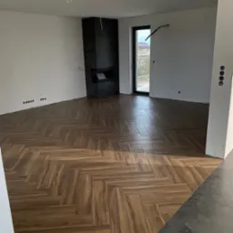 Ułożona podłoga z paneli w jodełkę w jasnym pomieszczeniu z kominkiem, ściany przygotowane do malowania, widoczne otwory na gniazdka.