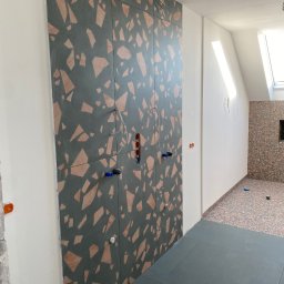 CER-MUR Przemysław Rosik - Ściana wyłożona panelami z wzorem lastryko w odcieniach szarości i brązu, z widocznymi instalacjami hydraulicznymi i elektrycznymi w trakcie montażu w pomieszczeniu z poddaszem.