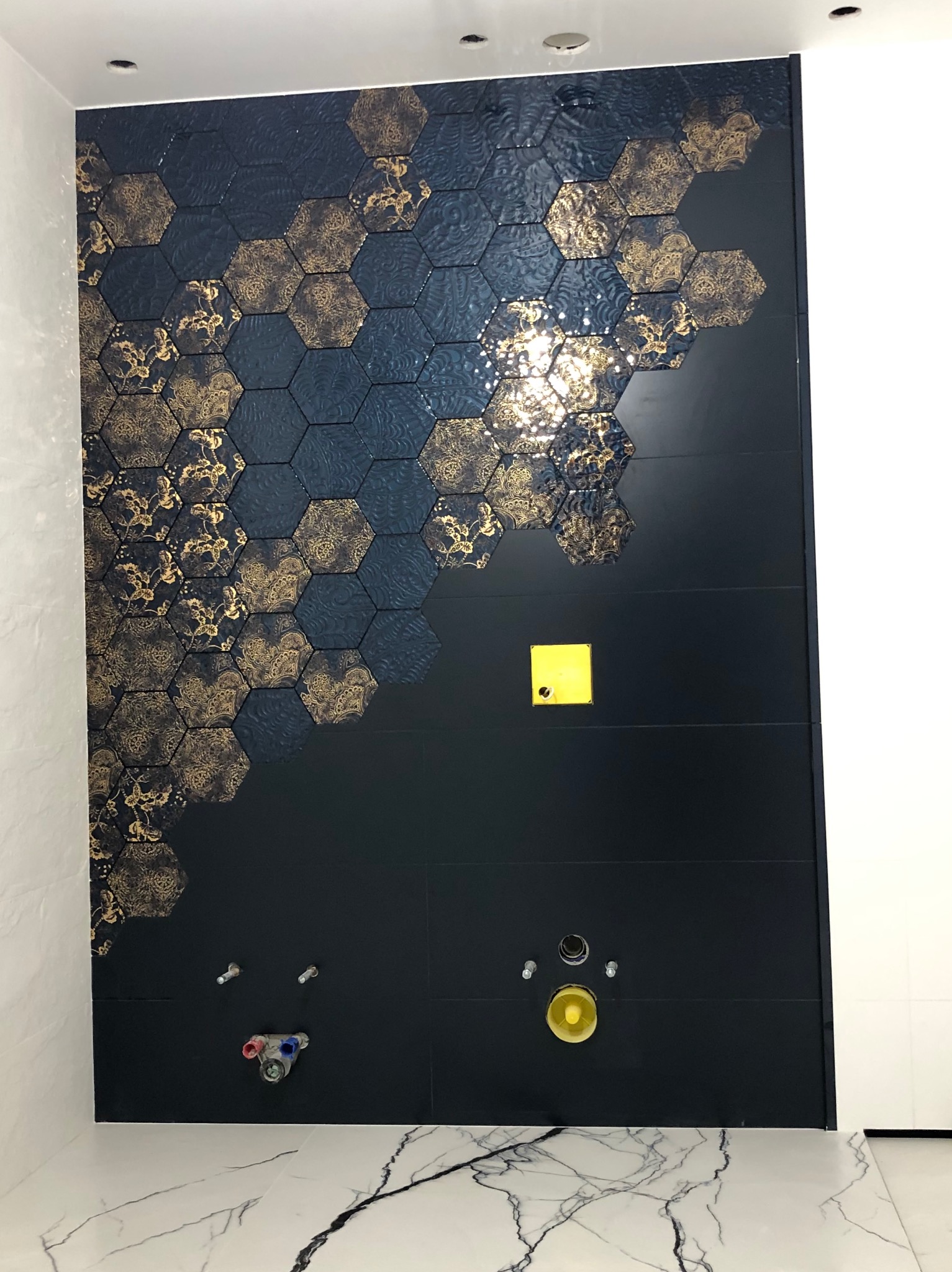 Ściana łazienki wyłożona ciemnymi płytkami heksagonalnymi z motywem złotych ornamentów, widoczne przyłącza wodne i kanalizacyjne, marmurowa posadzka.