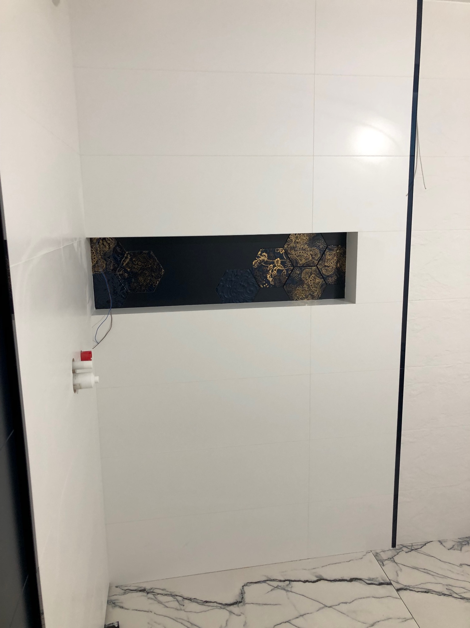 Białe płytki ścienne z wnęką zawierającą dekoracyjne, ciemne heksagonalne płytki ze złotym wzorem; marmurowa podłoga.