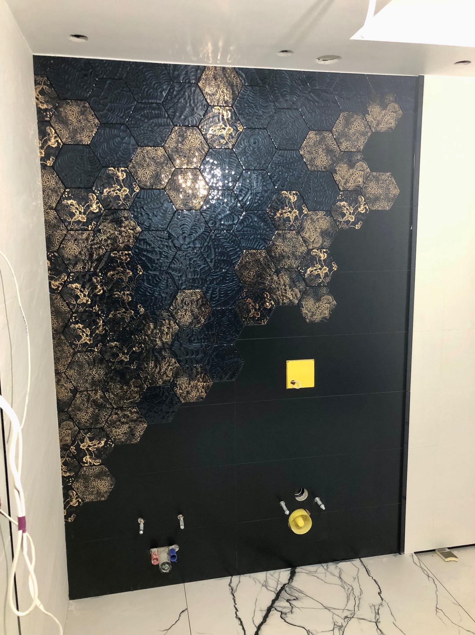 Ściana łazienki wykończona czarnymi heksagonalnymi płytkami z dekoracyjnym złotym wzorem, widoczne przyłącza hydrauliczne, fragment marmurowej podłogi.