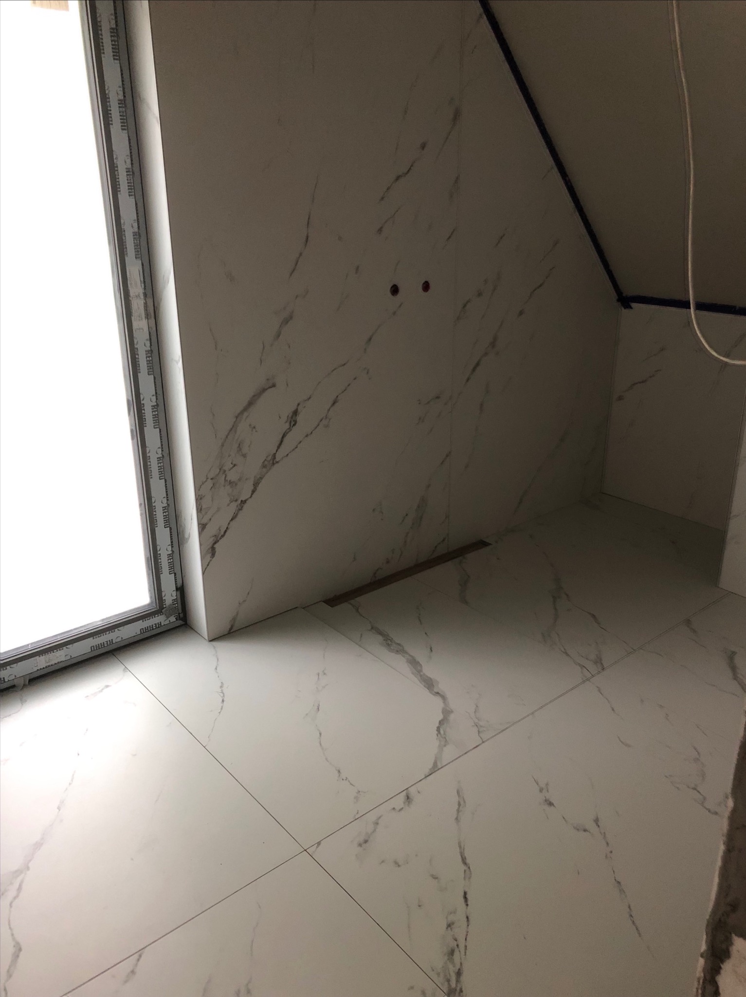 Wykończona łazienka w poddaszu z białymi płytkami imitującymi marmur na podłodze i ścianach, widoczne przyłącza do baterii oraz odpływ liniowy.