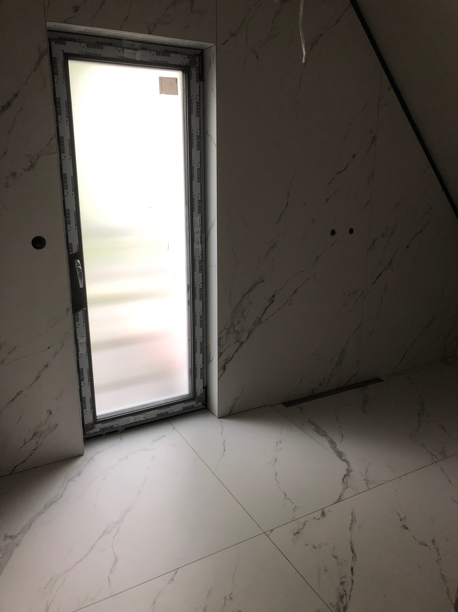 Nowoczesna łazienka na poddaszu z marmurowymi płytkami na ścianach i podłodze, okno z matową szybą w czarnej ramie, widoczne przygotowane otwory na instalacje.