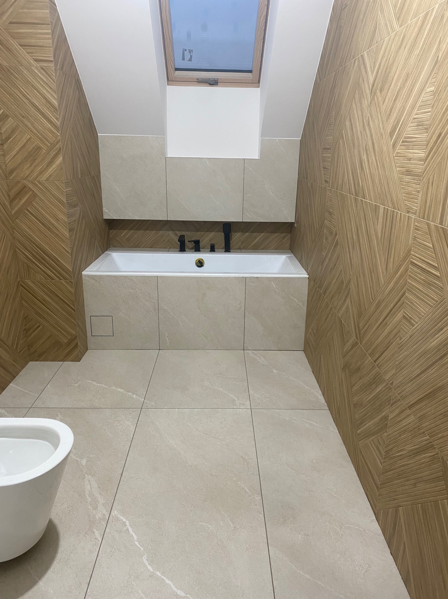 Nowoczesna łazienka na poddaszu z drewnopodobnymi płytkami na ścianach ułożonymi we wzór jodełki, beżowe płytki podłogowe imitujące kamień, biała wanna obudowana płytkami i czarna armatura, okno...