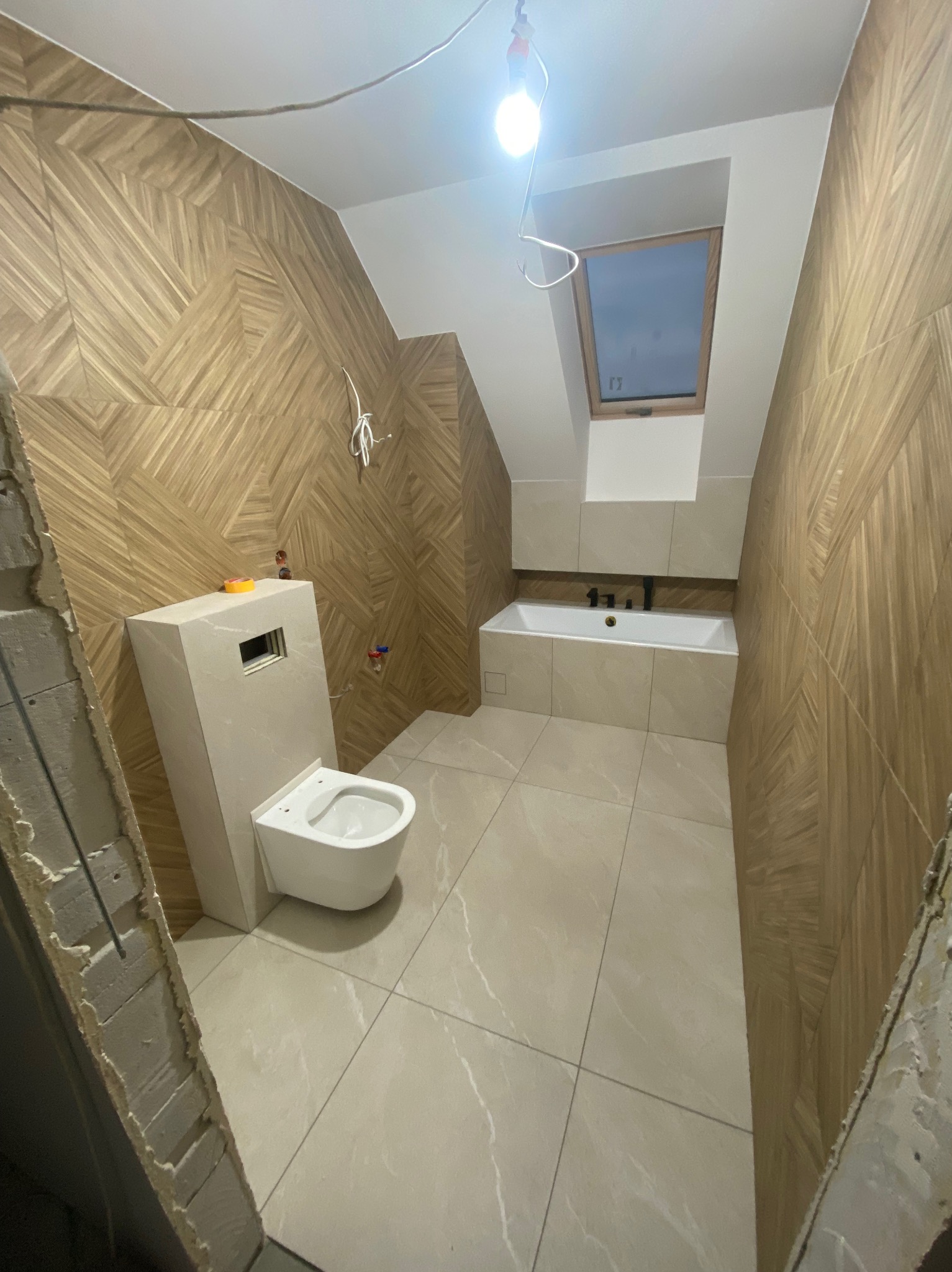 Wnętrze łazienki w trakcie remontu: drewnopodobne płytki ułożone w jodełkę na ścianach, zamontowana toaleta z zabudowaną spłuczką, wanna pod oknem dachowym, widoczne przewody i niedokończone...