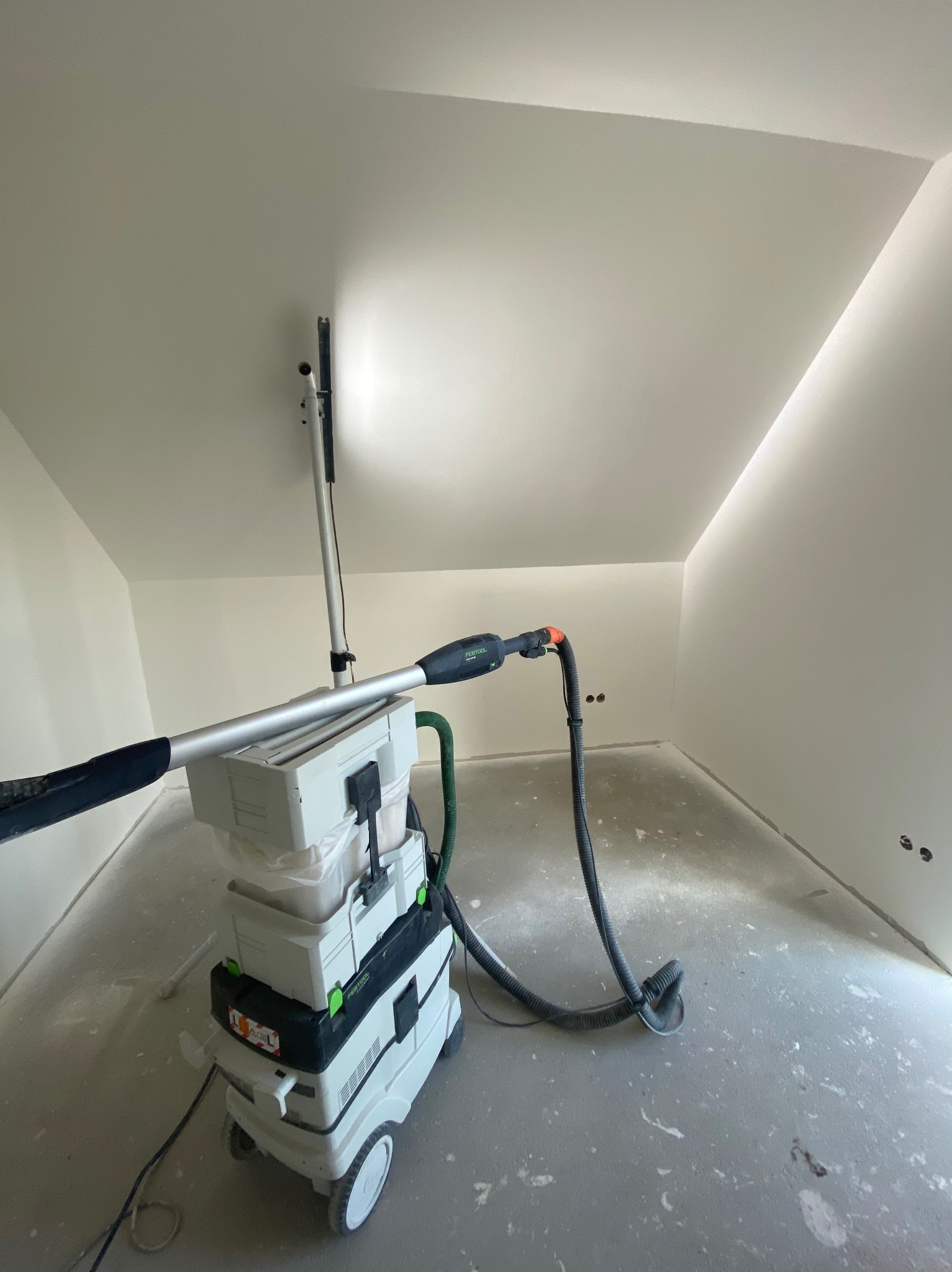 Szlifierka teleskopowa Festool Planex LHS 225 EQ z odkurzaczem podczas szlifowania sufitu w pomieszczeniu ze skosami, widoczne ślady pyłu na podłodze.