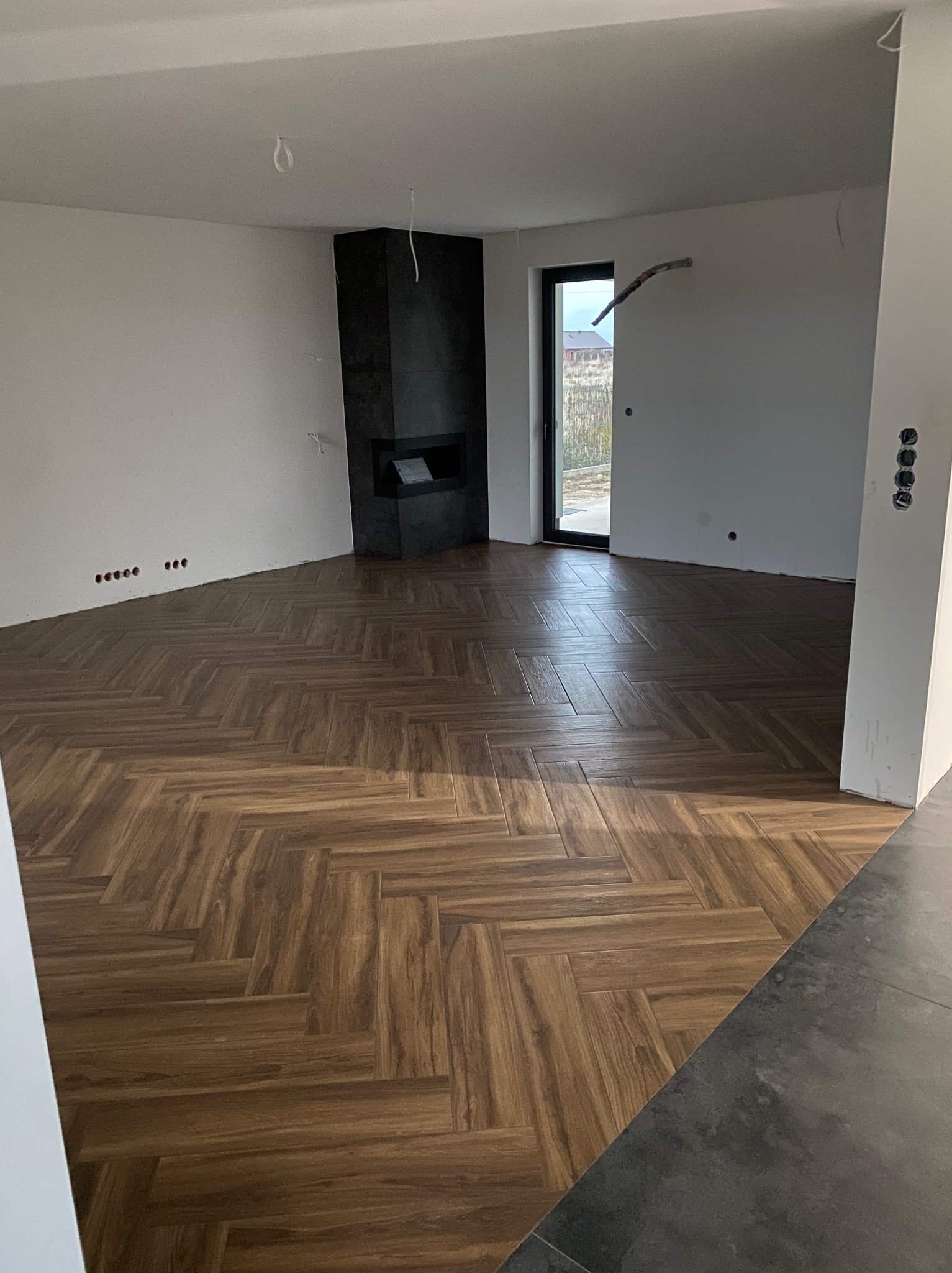 Ułożona podłoga z paneli w jodełkę w jasnym pomieszczeniu z kominkiem, ściany przygotowane do malowania, widoczne otwory na gniazdka.