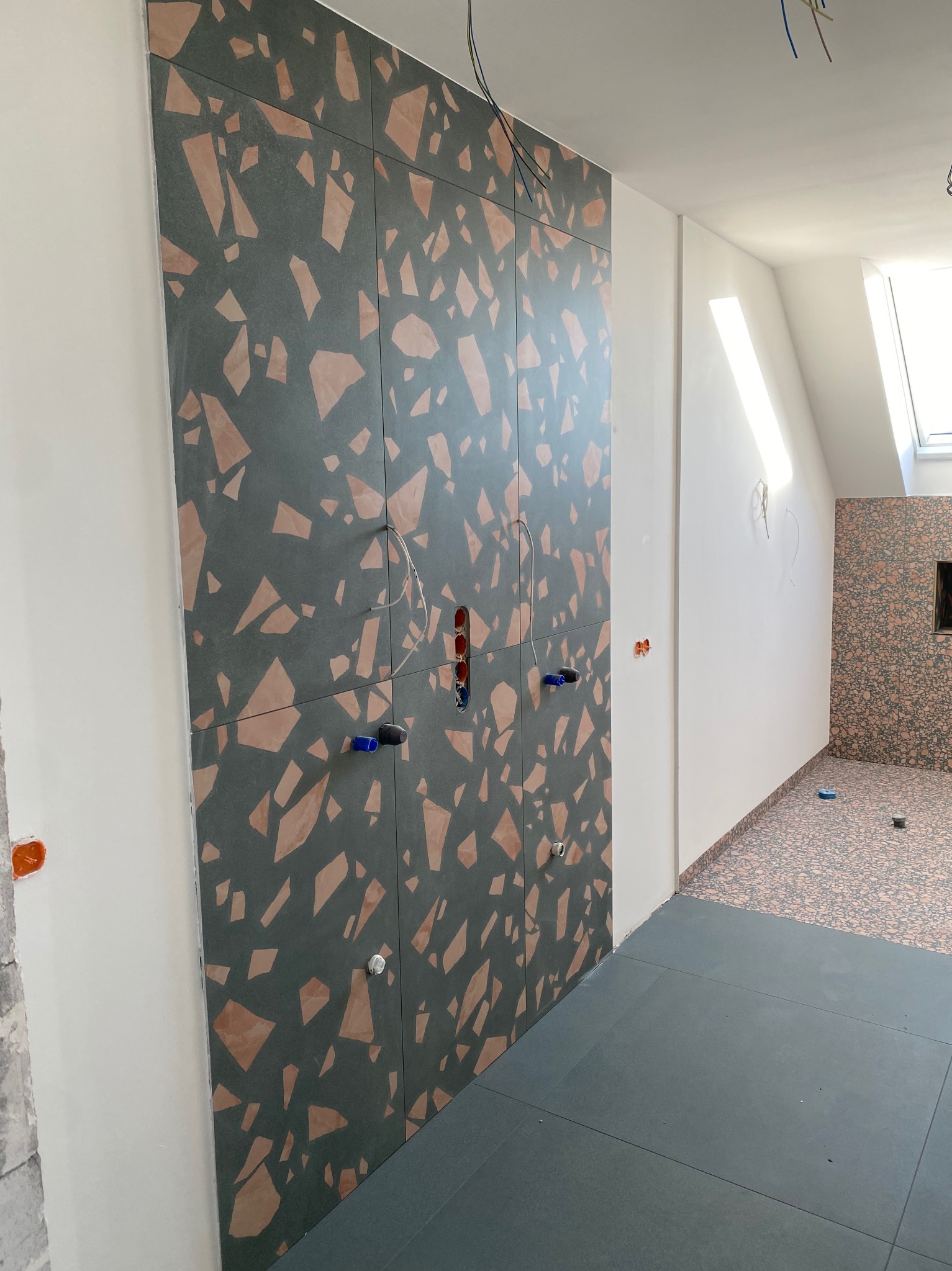 Ściana wyłożona panelami z wzorem lastryko w odcieniach szarości i brązu, z widocznymi instalacjami hydraulicznymi i elektrycznymi w trakcie montażu w pomieszczeniu z poddaszem.