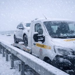 Holiday Łukasz Nowicki - Laweta z załadowanym samochodem osobowym, oba pokryte śniegiem, stoją na poboczu drogi podczas intensywnych opadów śniegu. Widoczny numer telefonu i napis 'Pomoc Drogowa'.