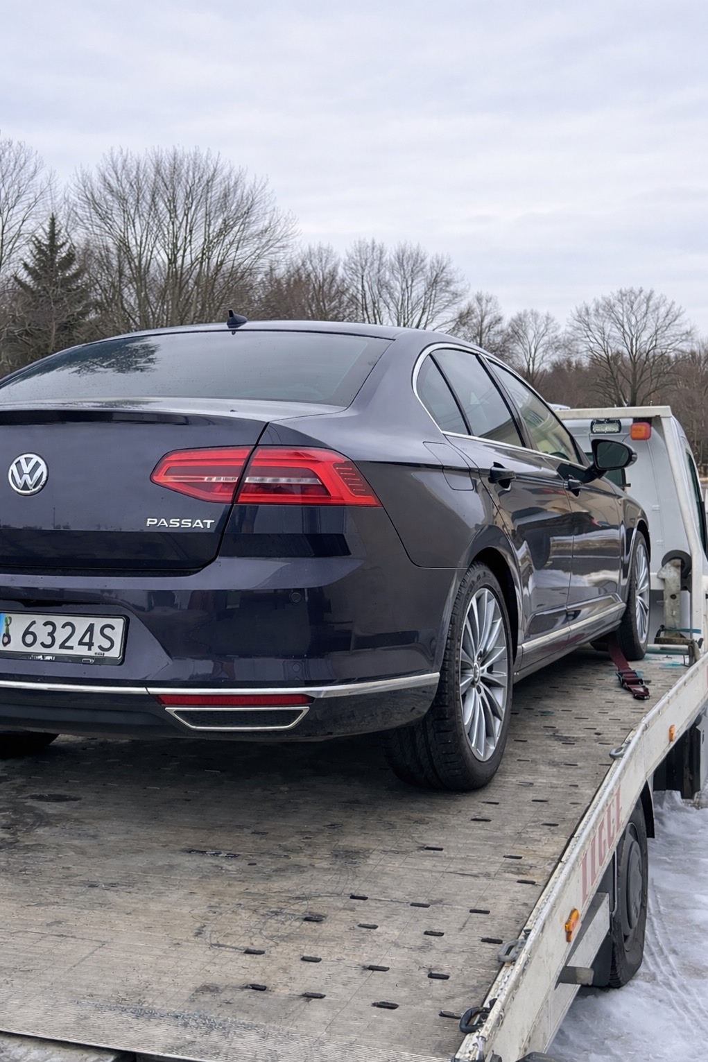 Granatowy Passat na lawecie w zimowy dzień. Widoczne tablice rejestracyjne i fragmenty drzew w tle. Ujęcie z tyłu pojazdu.