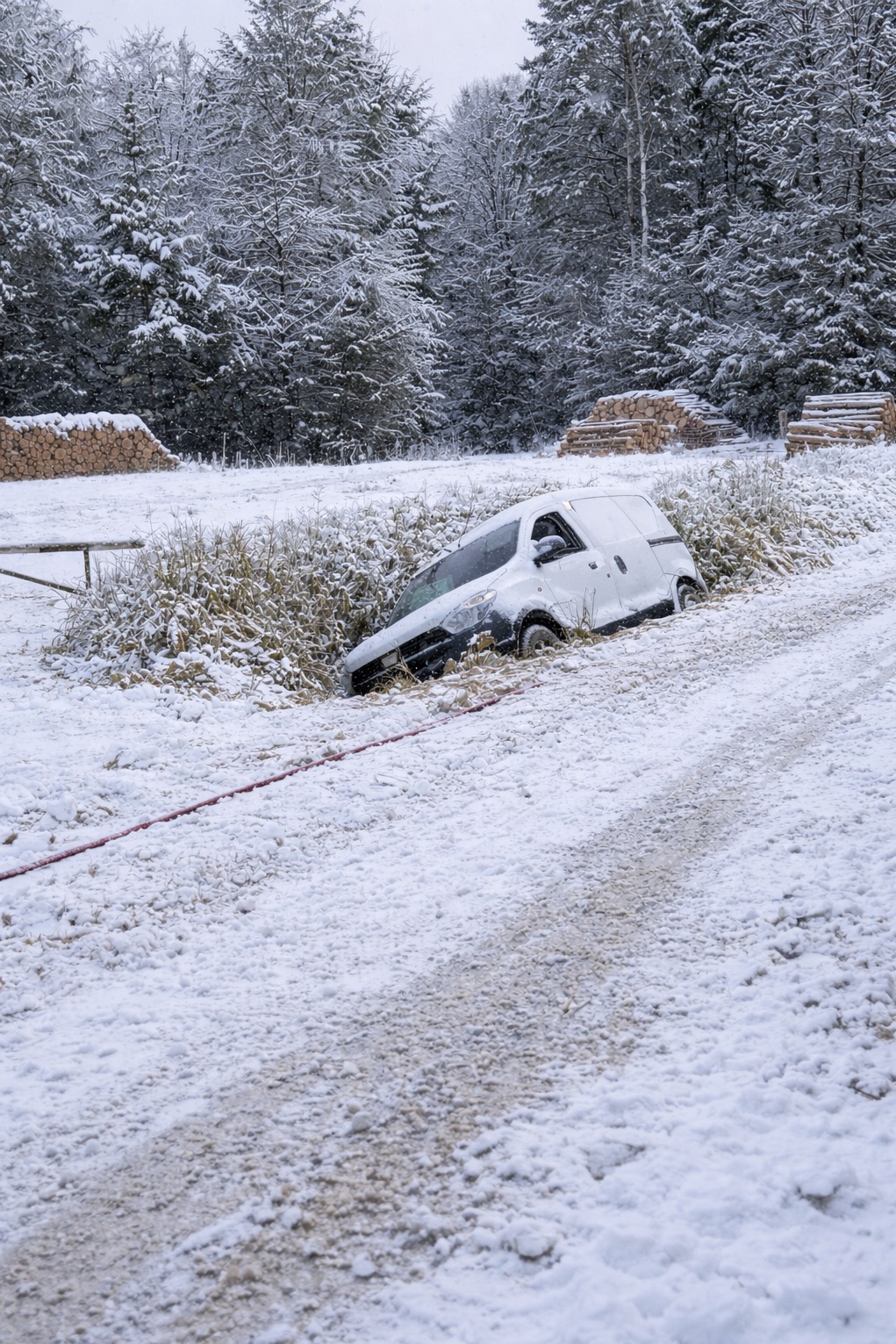 Zasypany śniegiem biały van w rowie na tle zimowego lasu. Czerwona lina holownicza widoczna na śniegu. Stosy drewna w tle.