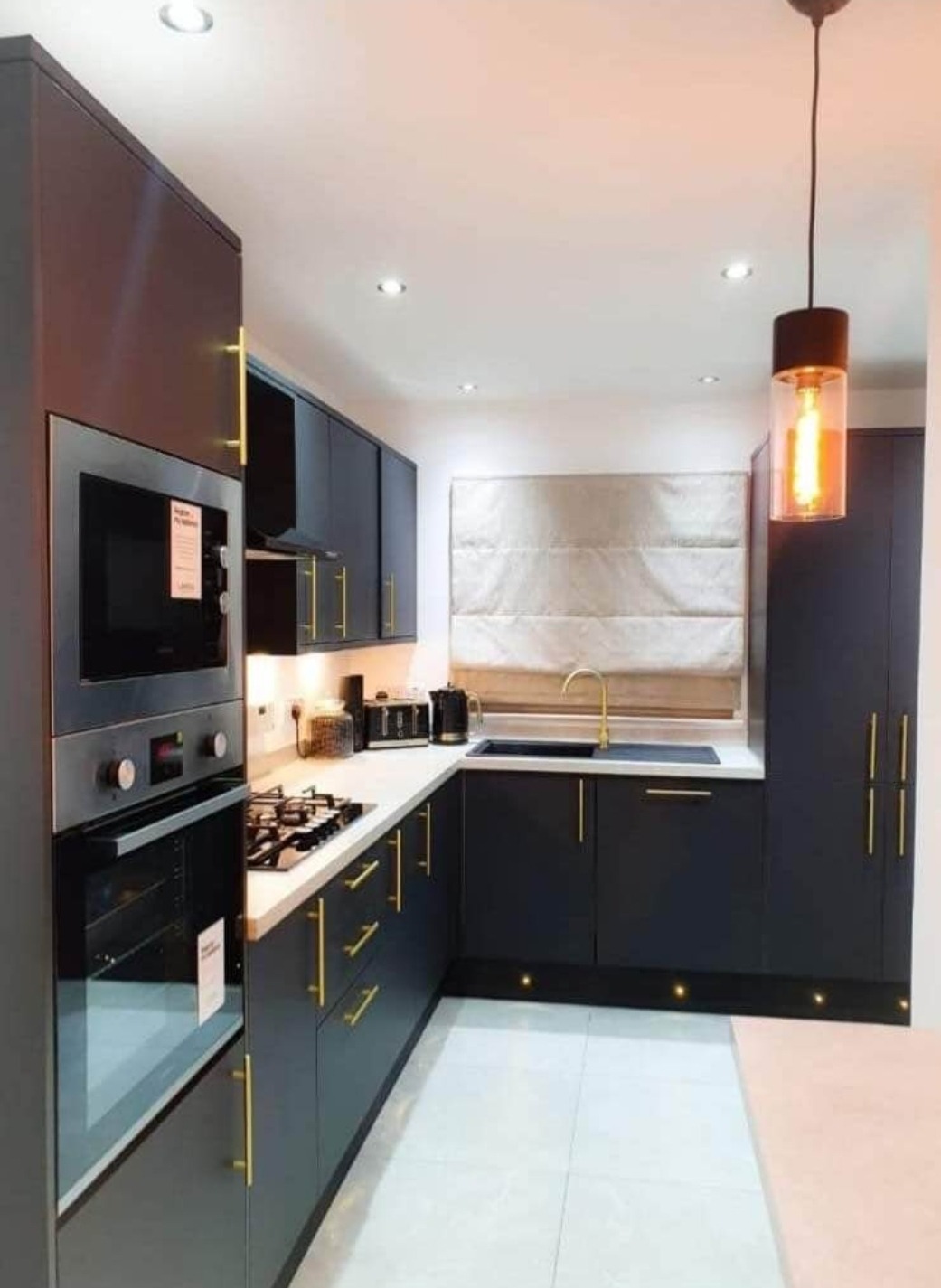 Nowoczesna kuchnia w odcieniach grafitu z białym blatem, złotymi uchwytami i armaturą, oświetlona wiszącą lampą z dekoracyjną żarówką i podświetleniem pod szafkami.