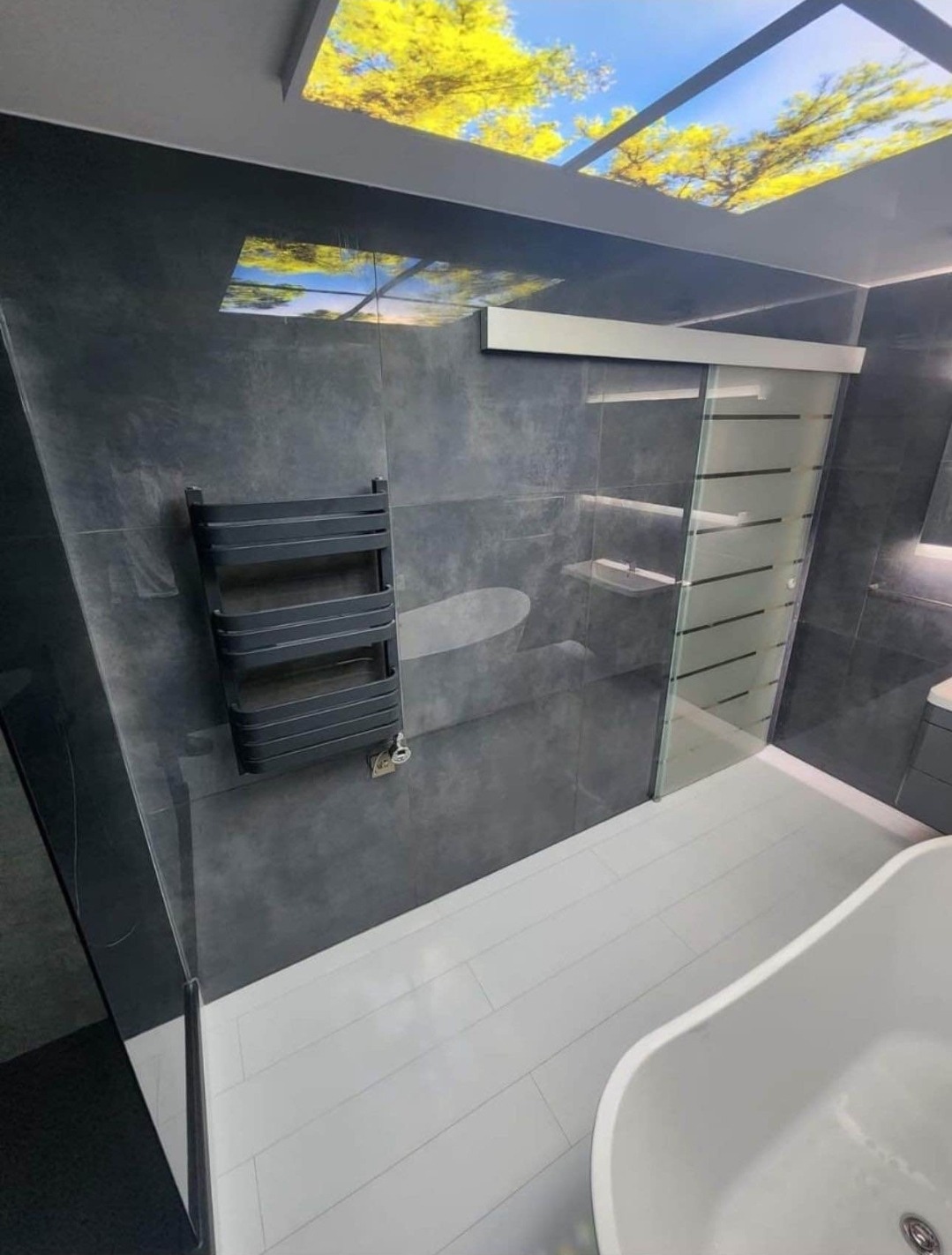 Nowoczesna łazienka z szarymi płytkami, białą podłogą imitującą deski i podświetlanym sufitem z motywem drzew, widoczny grzejnik drabinkowy i przesuwne szklane drzwi kabiny prysznicowej.