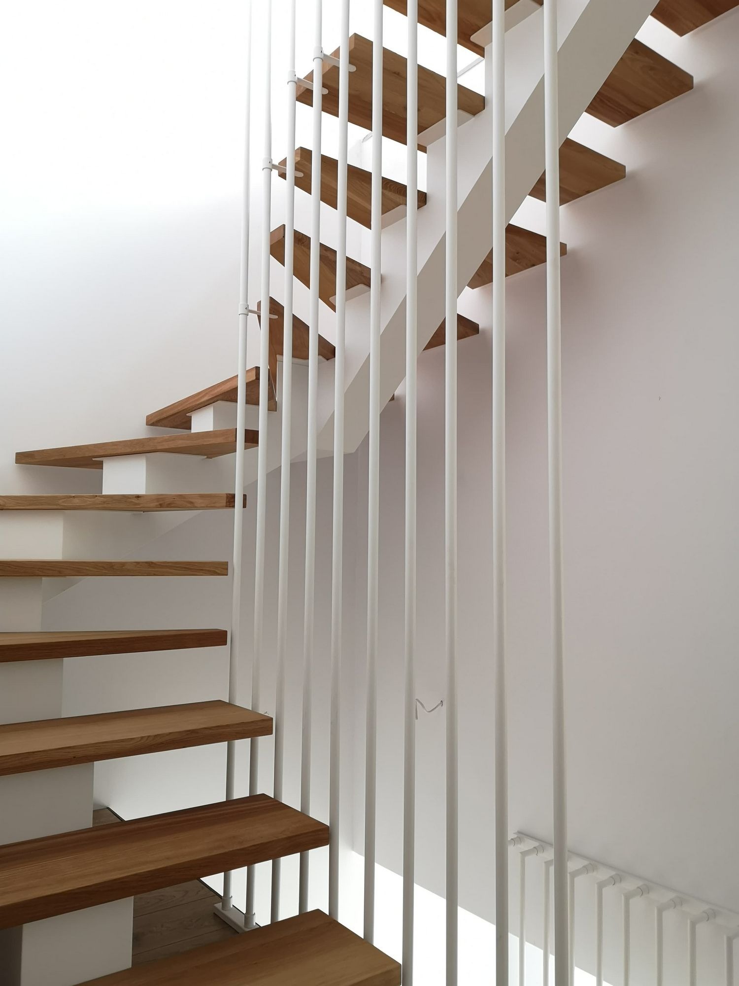 Minimalistyczne schody z drewnianymi stopniami i białymi, pionowymi balustradami w jasnym wnętrzu.