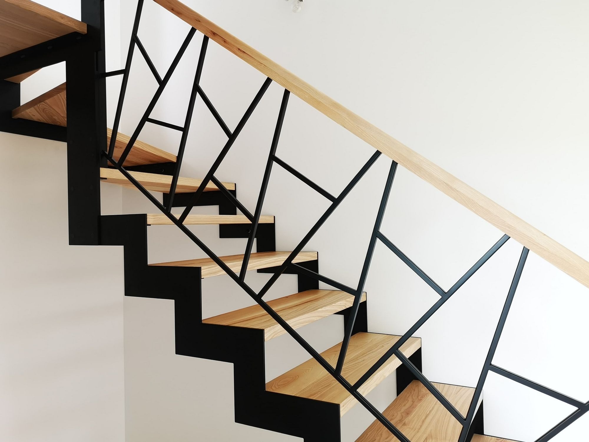 Nowoczesne schody metalowe z drewnianymi stopniami i geometryczną balustradą z czarnych profili, jasne drewno poręczy, minimalistyczne wnętrze.