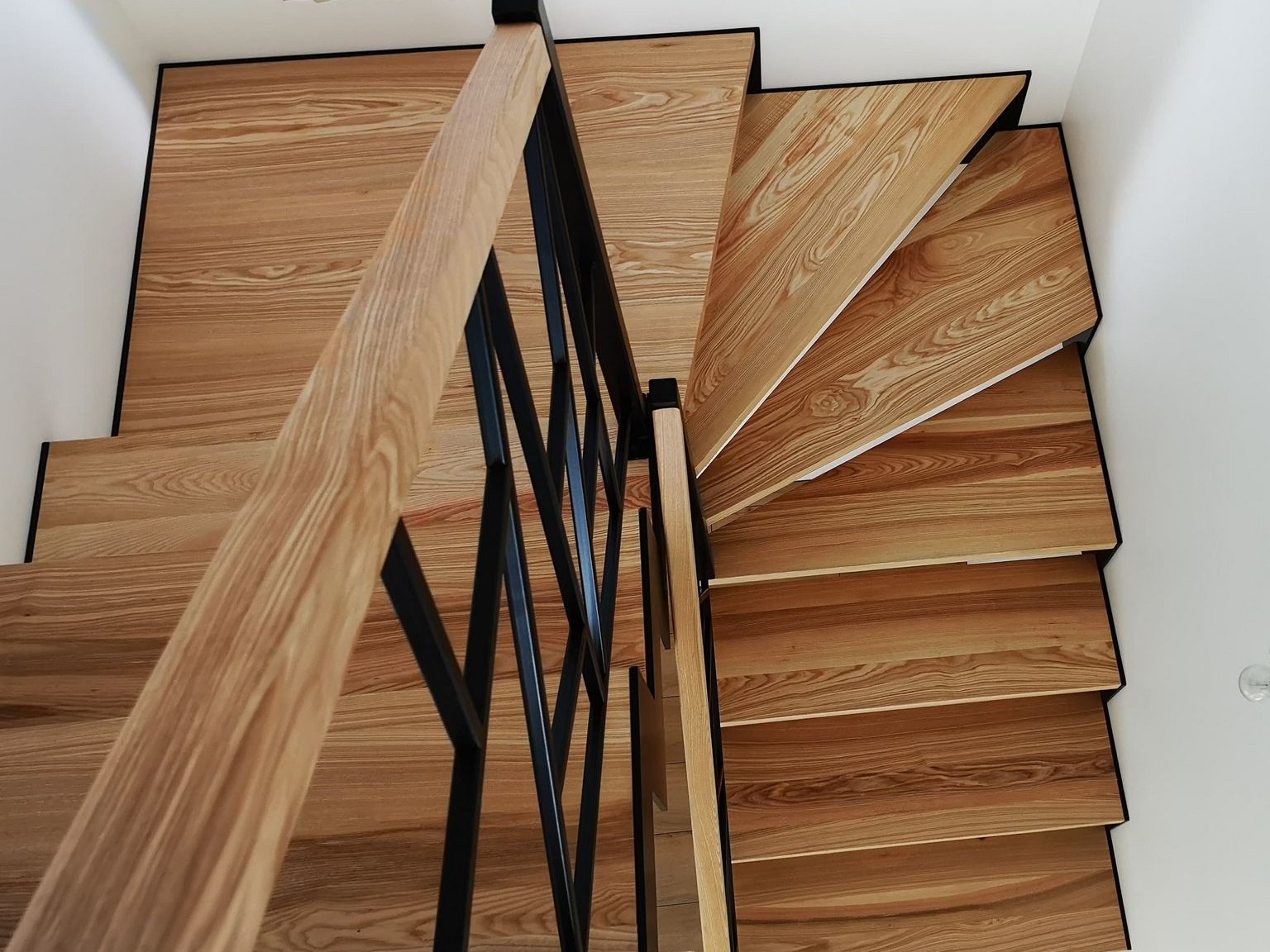 Klatka schodowa z drewnianymi stopniami o wyraźnym usłojeniu i czarną, geometryczną balustradą widziana z góry.