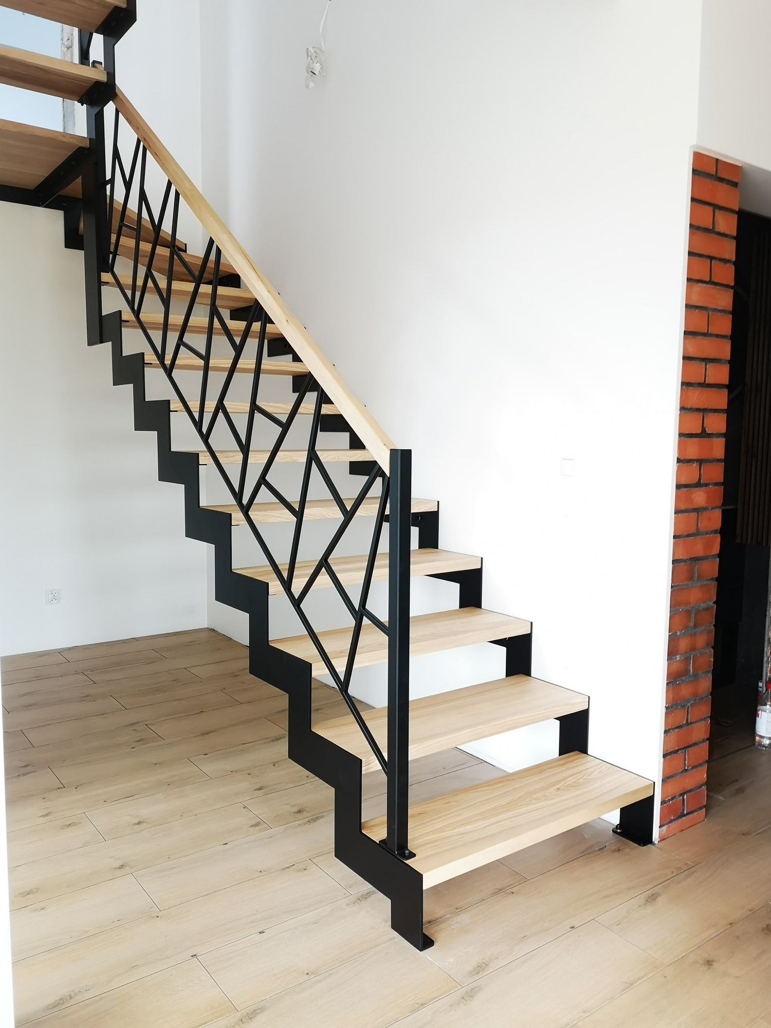 Nowoczesne schody wewnętrzne z drewnianymi stopniami i geometryczną, czarną metalową balustradą, widok od dołu, jasne wnętrze z parkietem i fragmentem ściany z cegły.
