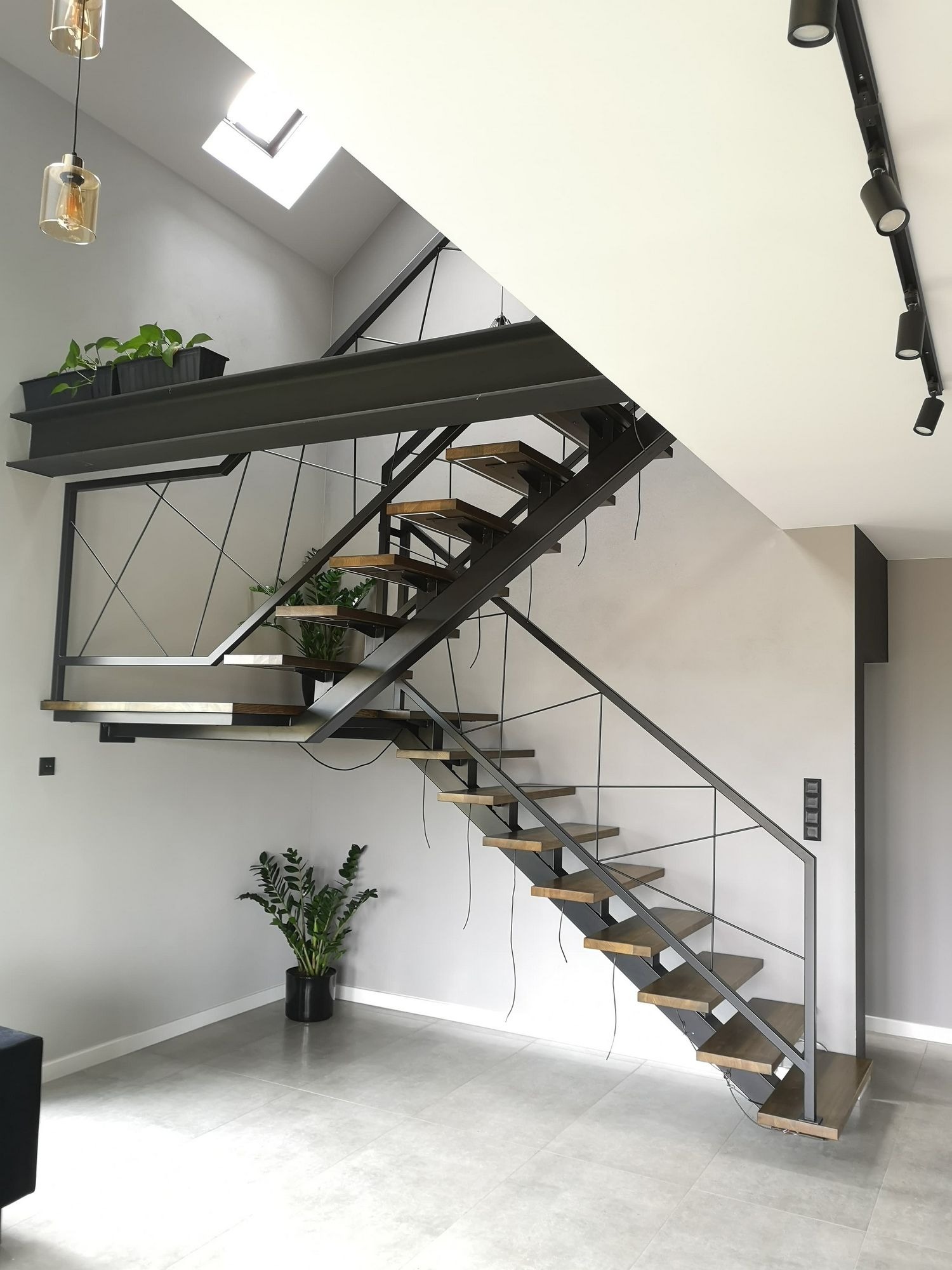 Nowoczesne schody z metalową konstrukcją i drewnianymi stopniami, oświetlone lampą wiszącą i reflektorami sufitowymi, z elementami dekoracyjnymi w postaci roślin doniczkowych.