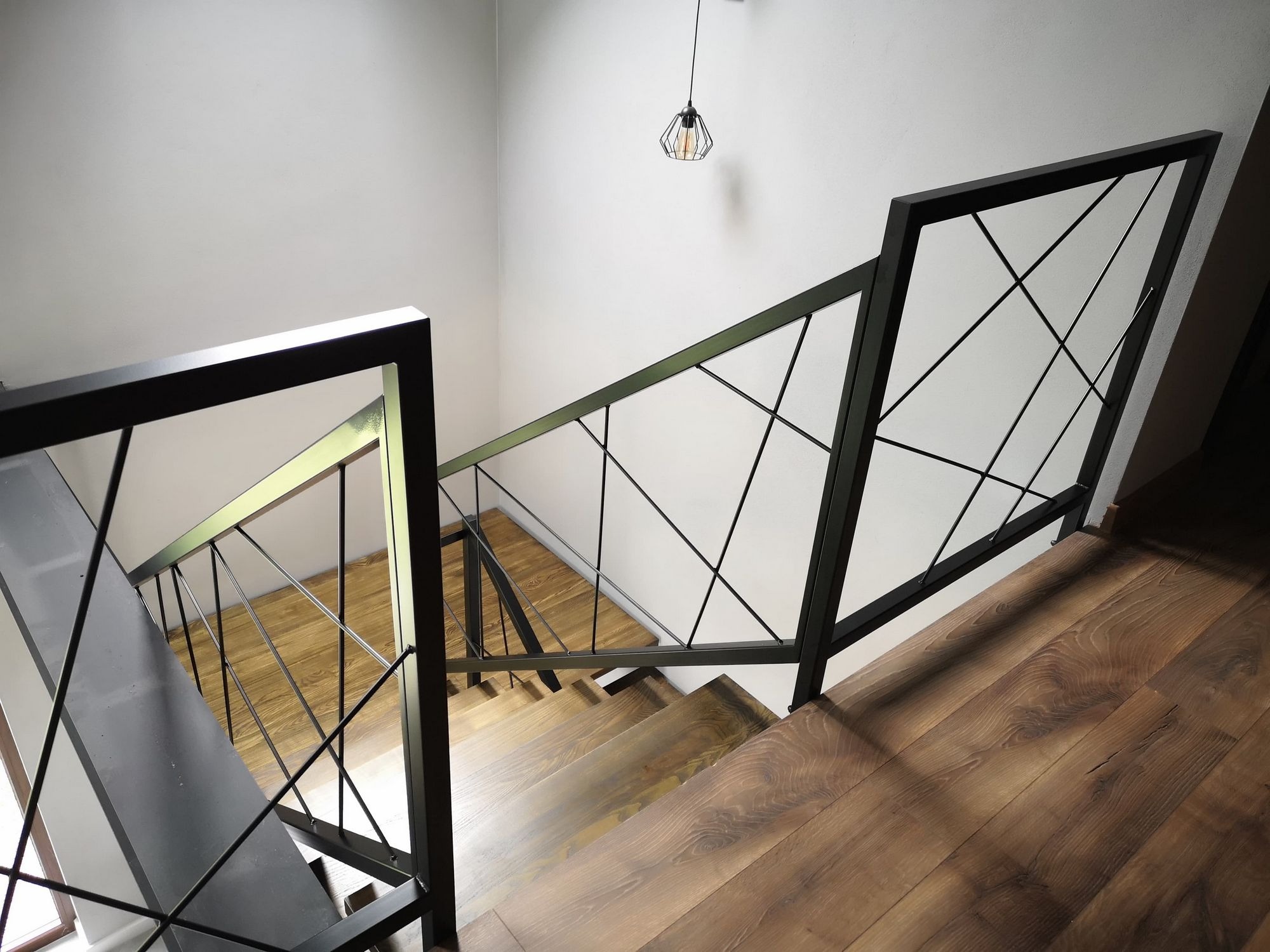Nowoczesne schody z drewnianymi stopniami i czarną, geometryczną balustradą metalową, widok z góry, wisząca lampa z metalową klatką.