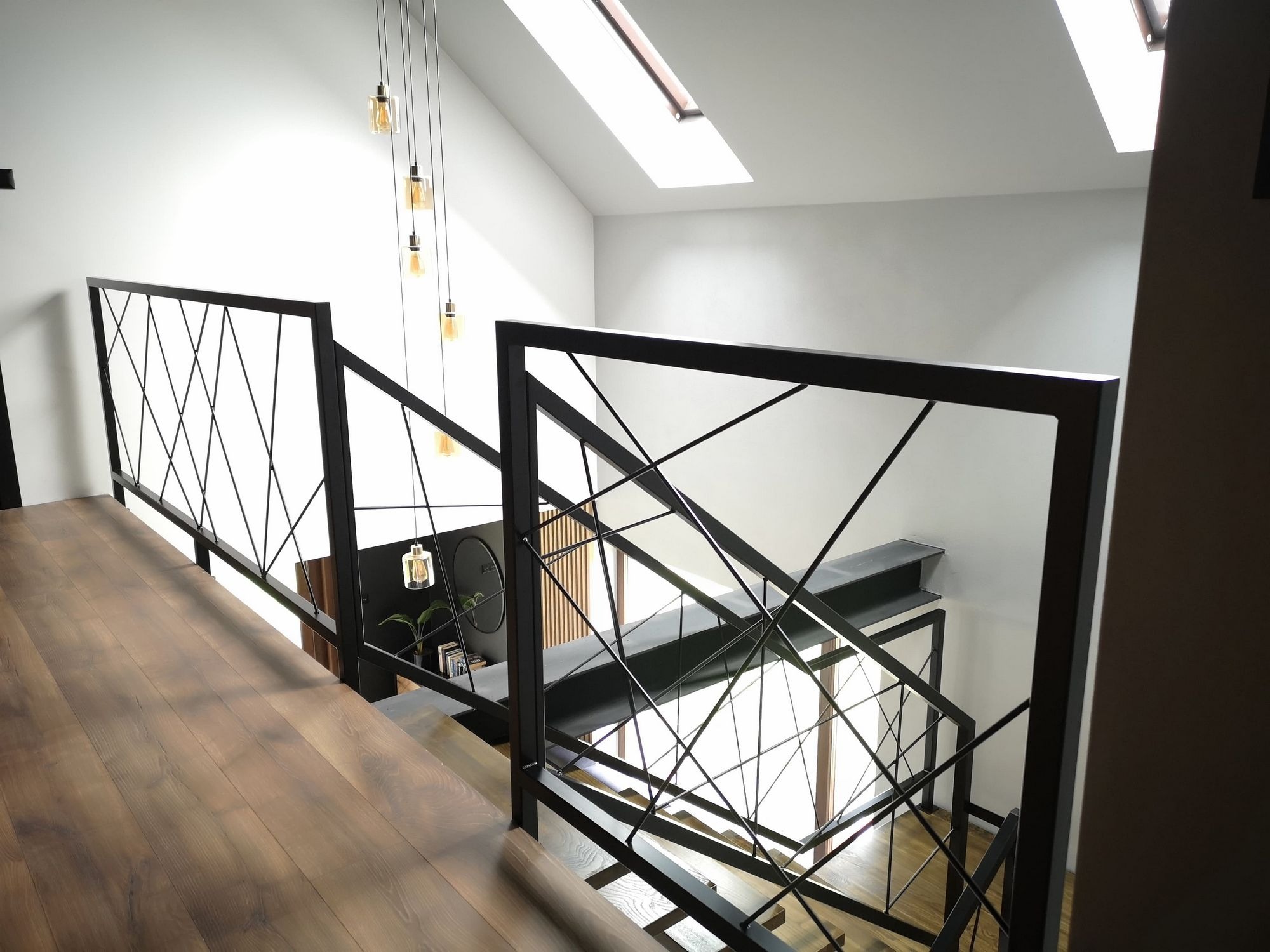 Nowoczesne schody z drewnianymi stopniami i metalową balustradą o geometrycznym wzorze, oświetlone wiszącymi lampami i światłem z okna dachowego.