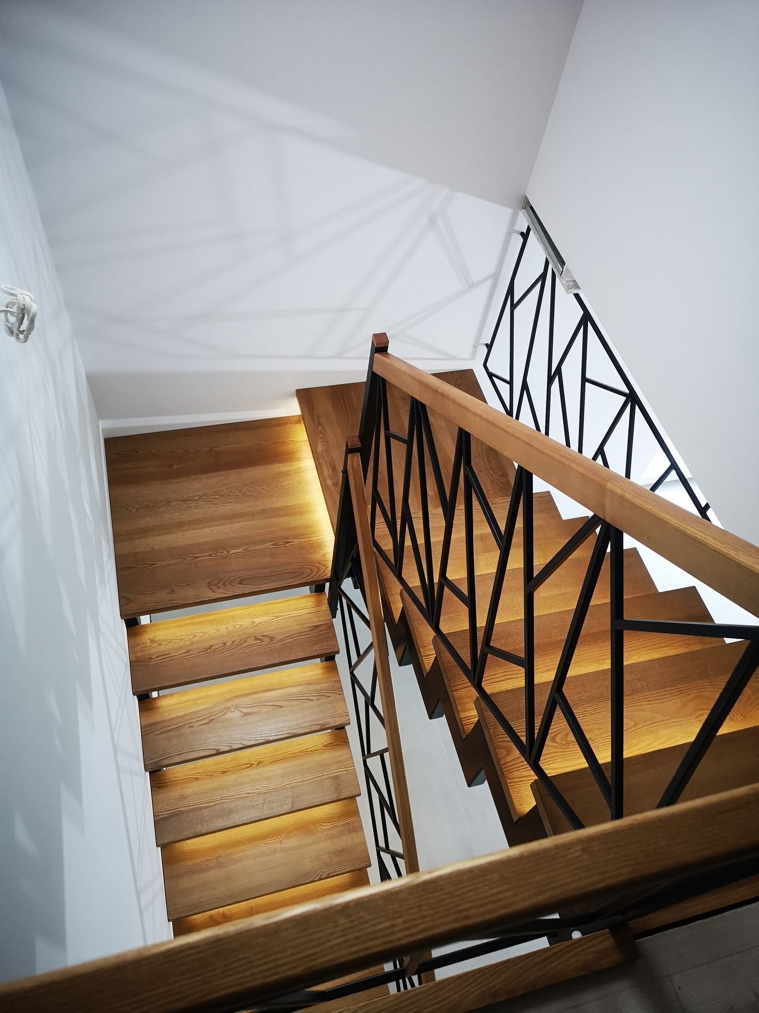 Nowoczesne schody z drewnianymi stopniami podświetlonymi od spodu i czarną, geometryczną balustradą, widok z góry.