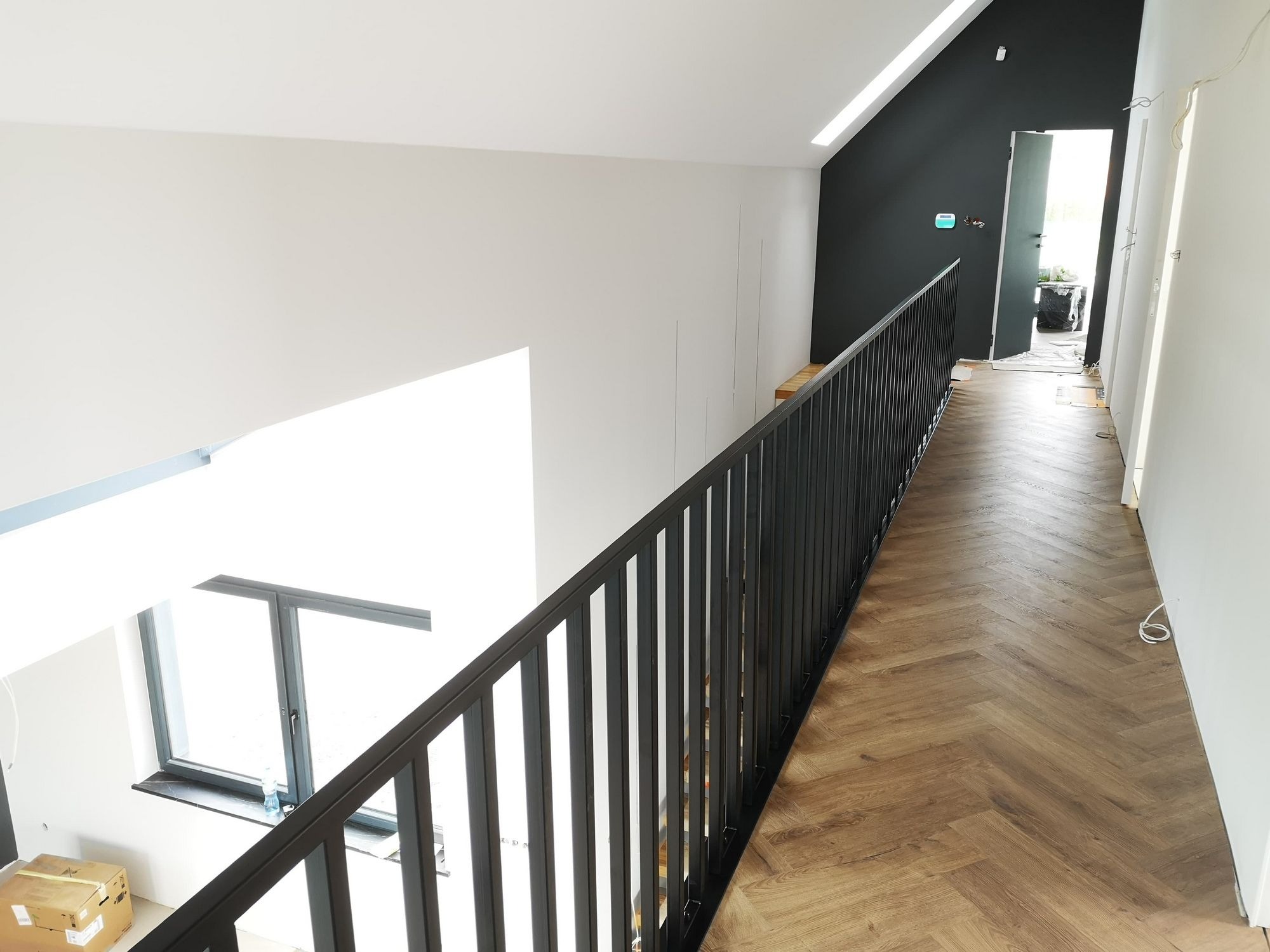 Nowoczesna czarna balustrada metalowa na antresoli z parkietem w jodełkę, widok z góry na okno i kartonowe pudła na podłodze.