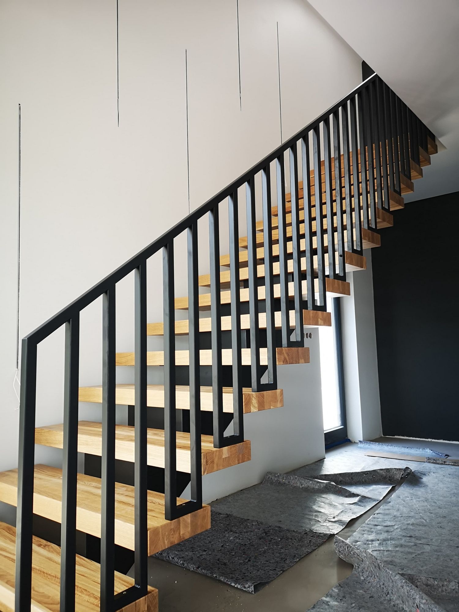 Nowoczesne schody z drewnianymi stopniami i czarną metalową balustradą, widoczne zabezpieczenia podłogi w trakcie montażu.
