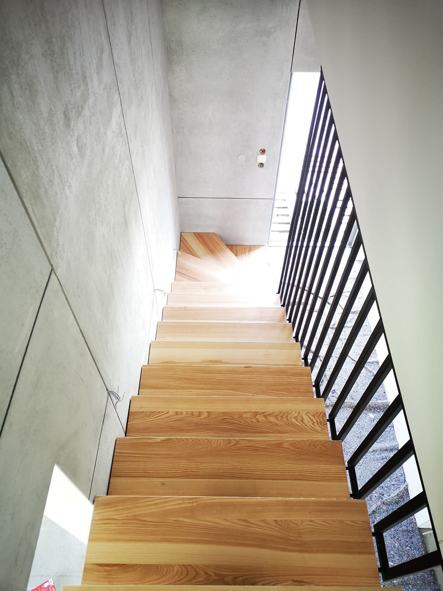 Nowoczesne schody z drewnianymi stopniami i metalową balustradą, widok od dołu w górę, jasne światło wpadające z góry, betonowa ściana po lewej stronie.