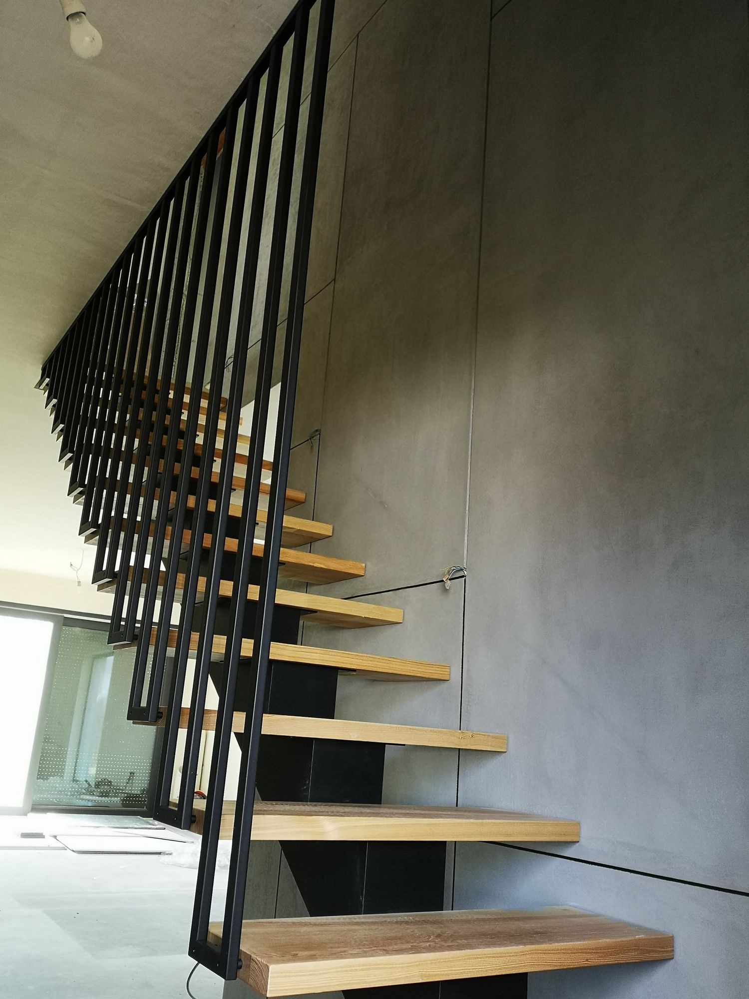 Nowoczesne schody z drewnianymi stopniami i czarną metalową balustradą w minimalistycznym wnętrzu z szarą ścianą.