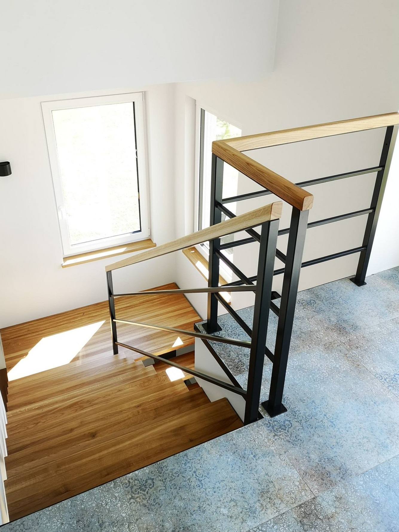 Nowoczesne schody z drewnianymi stopniami i czarną metalową balustradą z drewnianym poręczą, widok z góry na wzorzyste płytki podłogowe.