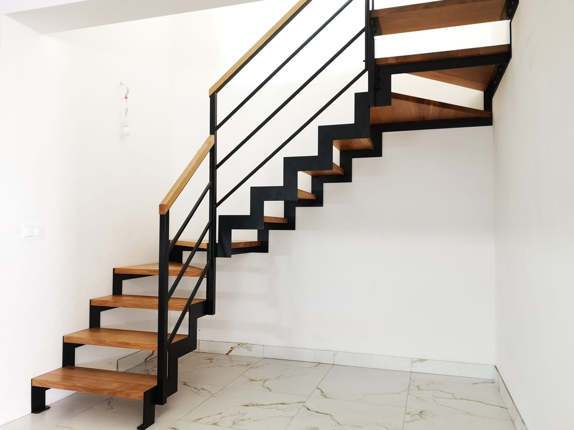 Nowoczesne schody z drewnianymi stopniami i czarną metalową konstrukcją oraz jasną drewnianą poręczą, na tle białych ścian i marmurowej podłogi.
