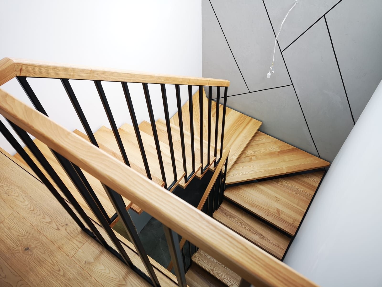 Kątowe ujęcie drewnianych schodów z czarnymi metalowymi balustradami, widok z góry, z nowoczesnym geometrycznym wzorem na ścianie w tle.
