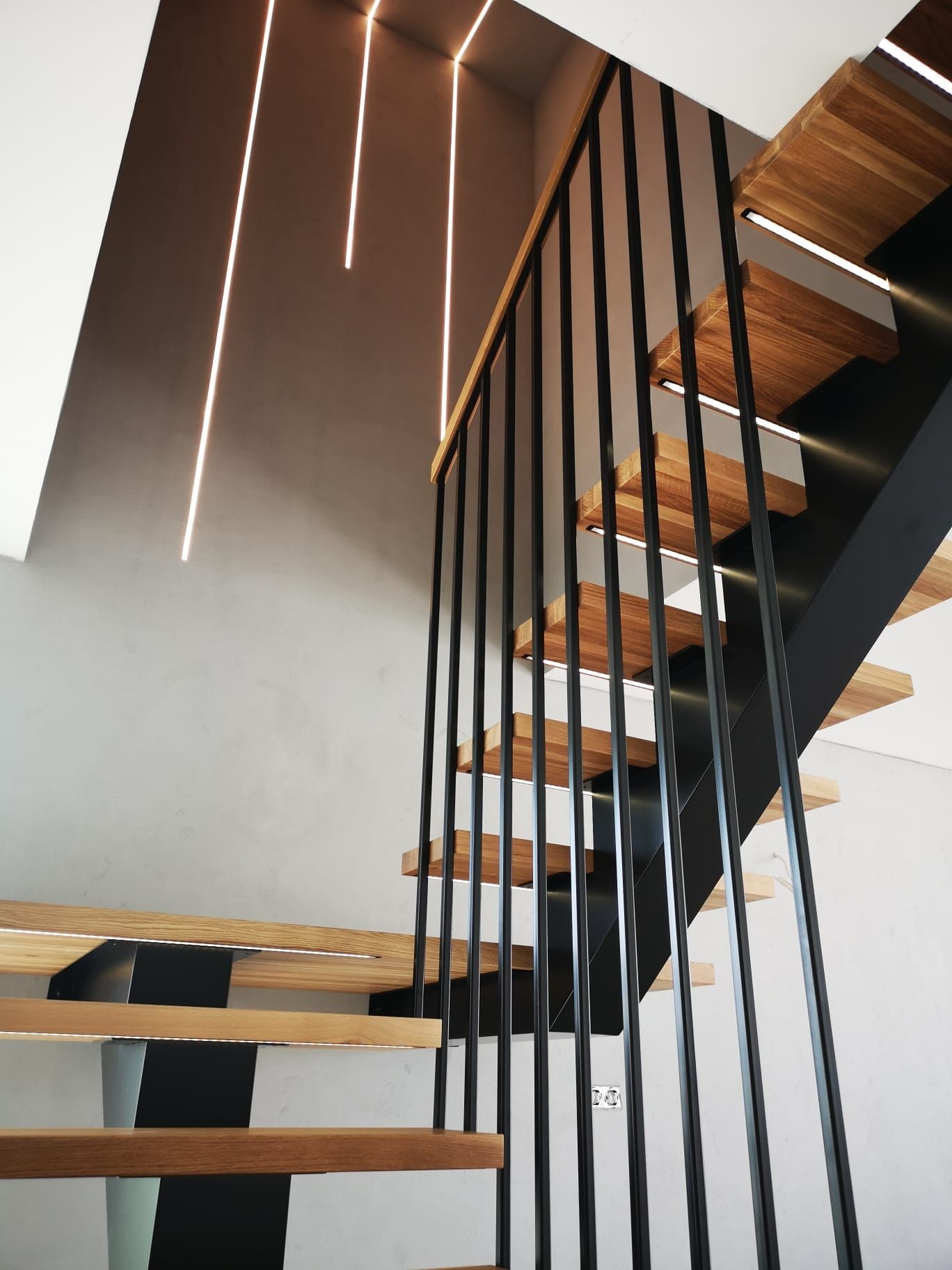 Nowoczesne schody z drewnianymi stopniami podświetlonymi od spodu i czarną metalową balustradą z pionowymi prętami, oświetlenie sufitowe w postaci pionowych linii świetlnych.
