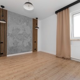 DC HOME SPÓŁKA Z OGRANICZONĄ ODPOWIEDZIALNOŚCIĄ - Pusty, jasny pokój z panelami ściennymi z listew drewnianych, szarą ścianą z efektem betonu, i lampami wiszącymi. Na podłodze panele podłogowe w odcieniach beżu i brązu. Okno z beżową zasłoną...