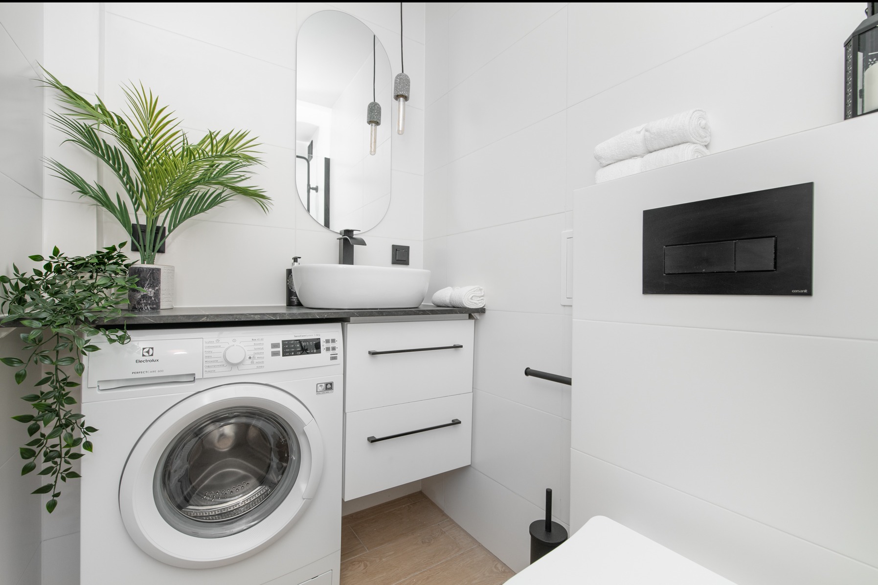 Nowoczesna, biała łazienka z pralką Electrolux PerfectCare 600, owalną umywalką nablatową, minimalistycznym lustrem i drewnopodobną podłogą.