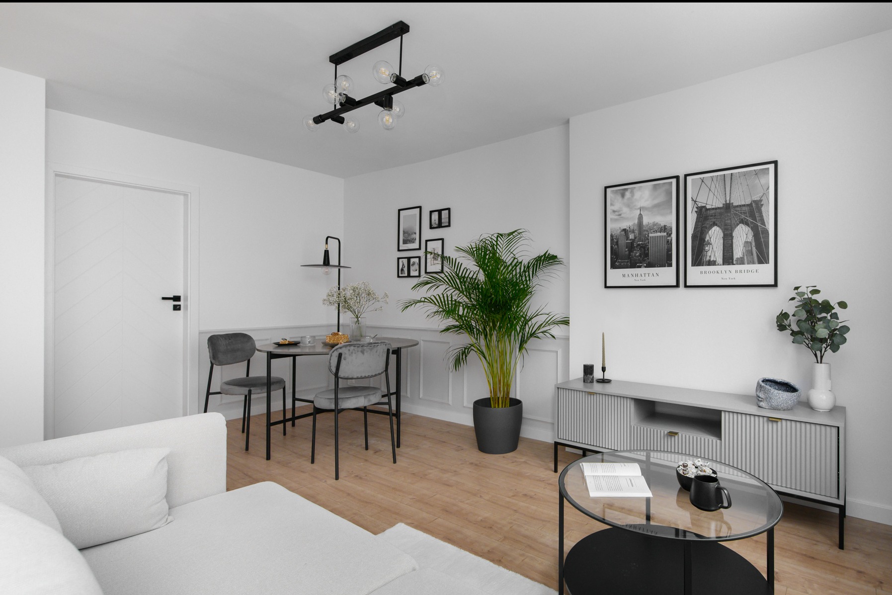 Jasne wnętrze salonu w stylu minimalistycznym z drewnianą podłogą, białymi ścianami z dekoracyjną sztukaterią, szarymi meblami, okrągłym stołem z krzesłami, roślinami w donicach i czarno-białymi...