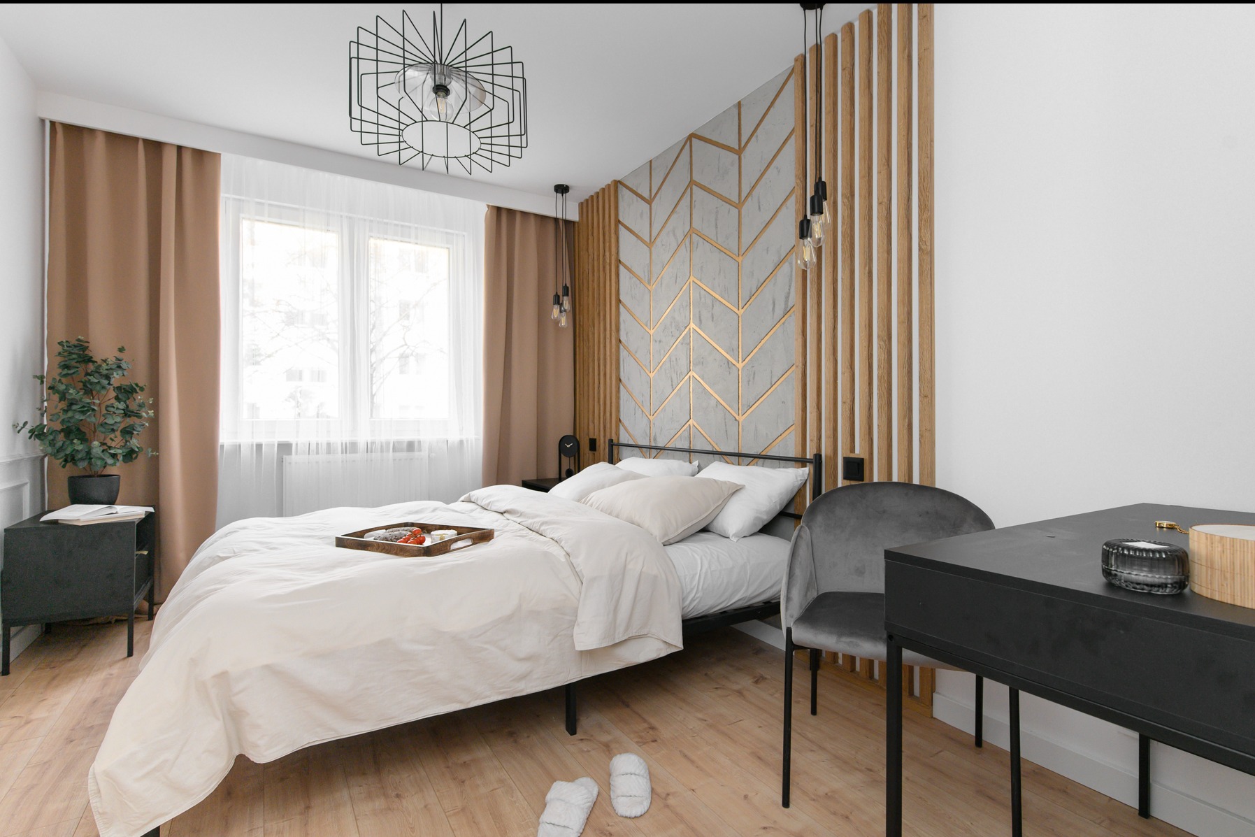 Nowoczesna sypialnia z łóżkiem, ścianą dekoracyjną z motywem jodełki i drewnianymi panelami, designerską lampą sufitową, biurkiem i szarą aksamitną krzesłem, na podłodze leżą białe kapcie.