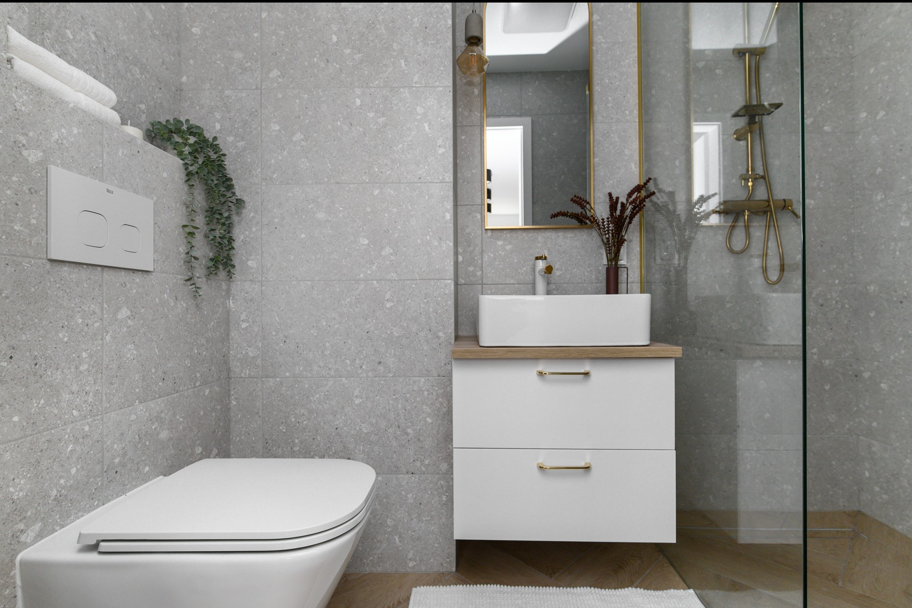 Nowoczesna łazienka w szarościach z wiszącą toaletą, umywalką nablatową na białej szafce z drewnianym blatem, złotą armaturą i szklaną kabiną prysznicową.