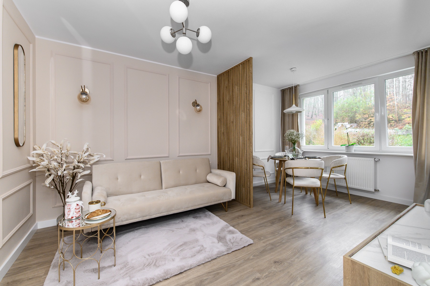 Jasne wnętrze mieszkania z beżową sofą, okrągłym stolikiem z przekąskami, dywanem imitującym marmur, drewnianą podłogą, ścianą z dekoracyjnymi panelami i wydzieloną częścią jadalną z widokiem...