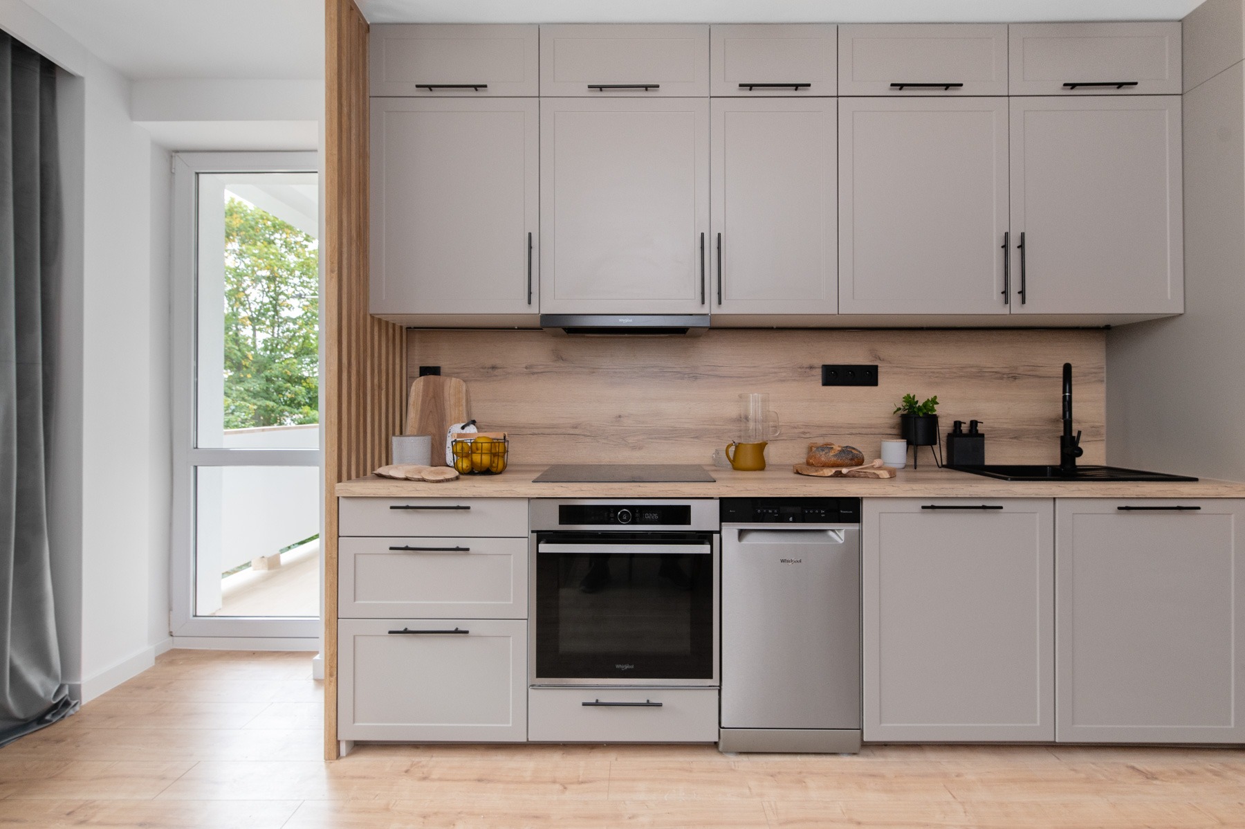 Jasnoszara kuchnia z drewnianym blatem i panelem, czarne uchwyty i armatura, okno z widokiem na zieleń, nowoczesny design.