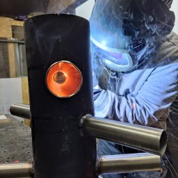 GMTK Sp. z o.o. - Spawacz w masce spawalniczej łączy metalowe rury do czarnego, pionowego elementu. Widoczne iskry i dym. Detal spoiny i otworu z pomarańczowym światłem wewnątrz.