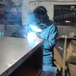 GMTK Sp. z o.o. - Spawacz w masce spawalniczej łączy metalowe elementy, iskry i dym w warsztacie. Widoczne narzędzia i elementy konstrukcji. Oświetlenie jarzeniowe.