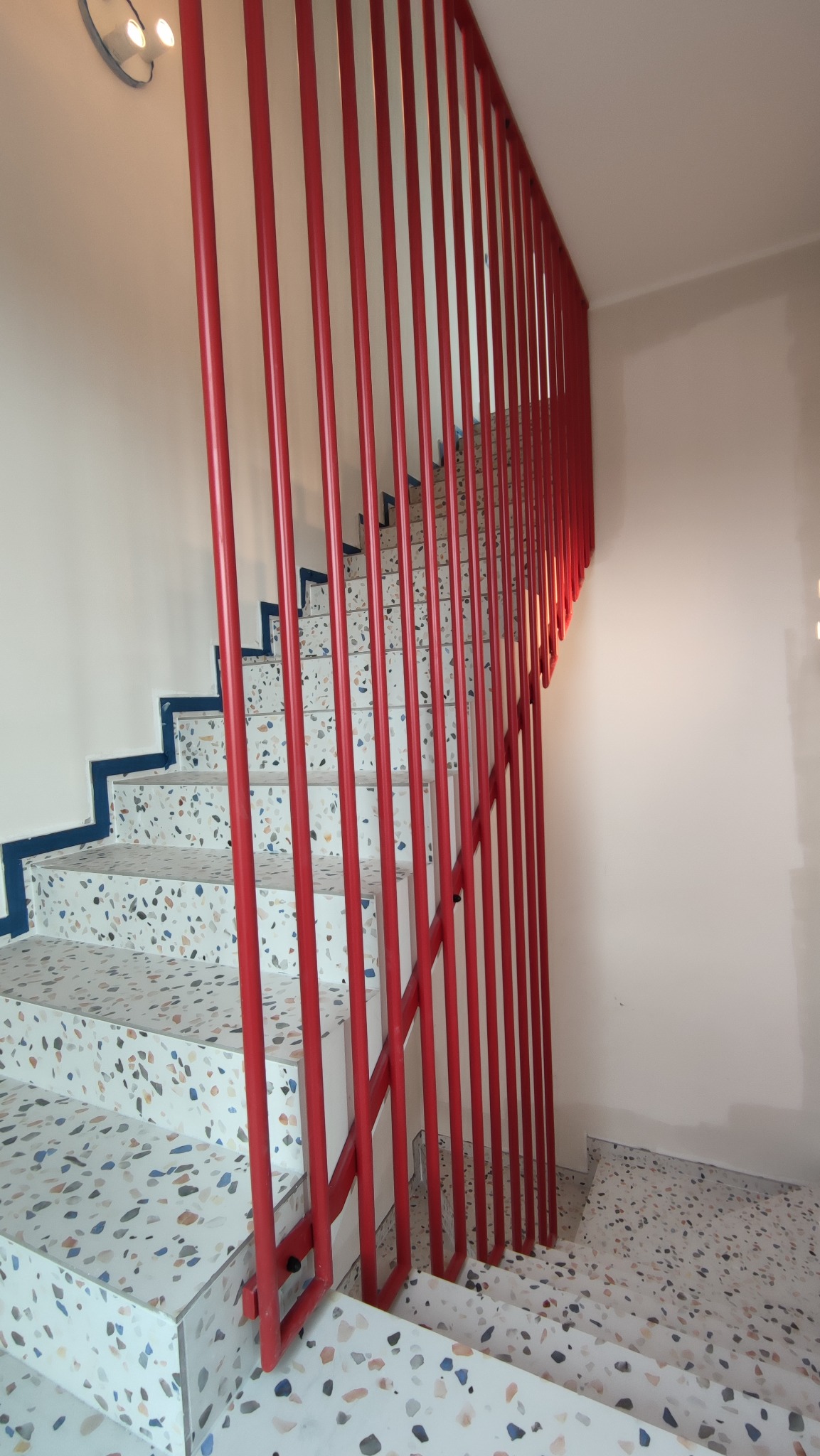 Wewnętrzne schody z terazzo z czerwoną, pionową balustradą. Ujęcie z dołu, ukazujące detale wykończenia i kolorystykę. Minimalistyczny design.