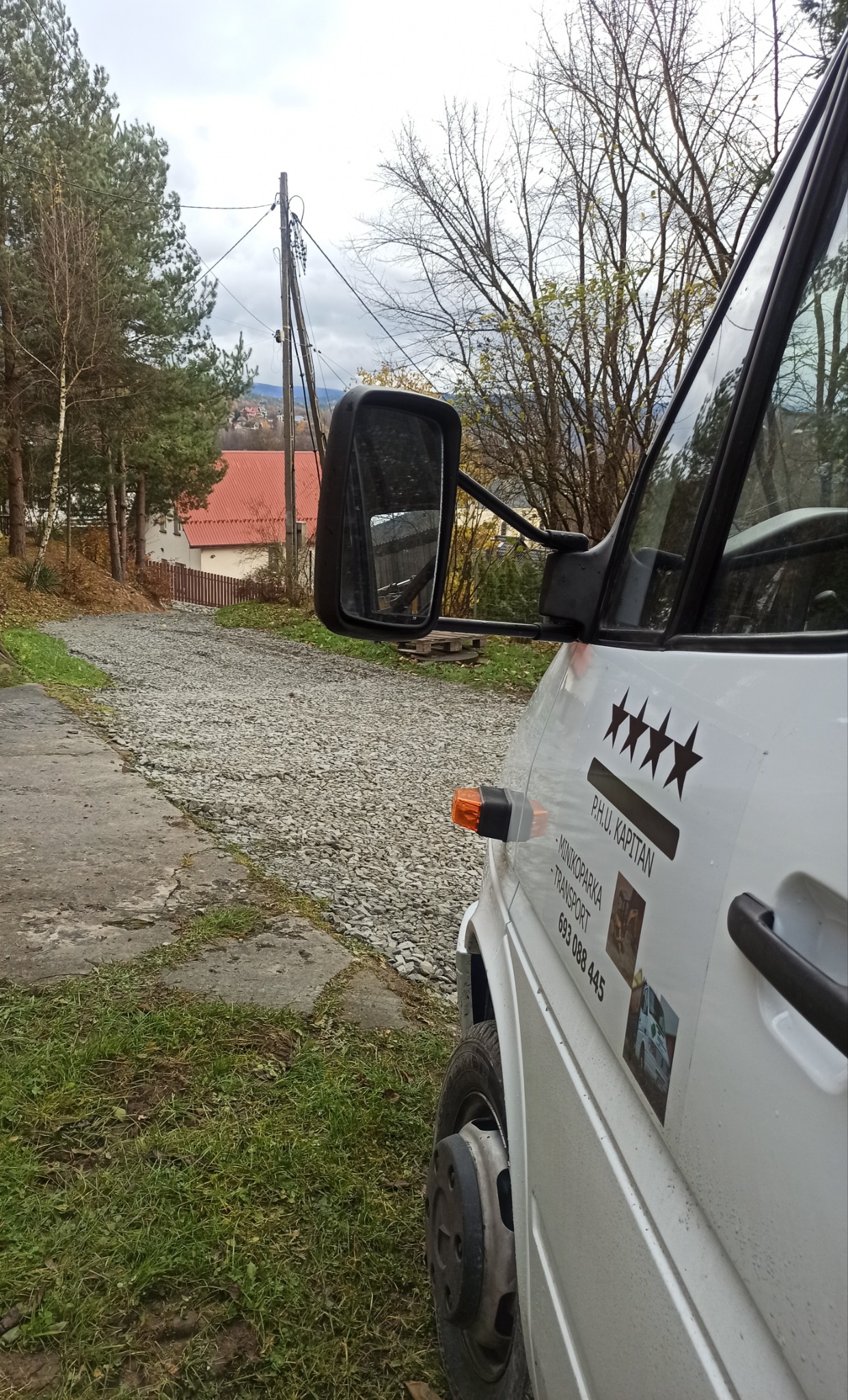 Widok z boku na biały samochód dostawczy z naklejkami firmy transportowej na tle drogi wysypanej żwirem, prowadzącej pod górę w kierunku zabudowań.