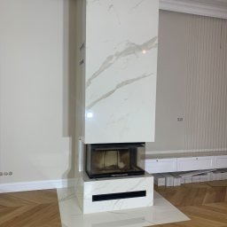 Grzegorz Zegzula - Nowoczesny kominek obudowany białym marmurem z subtelnym, szarym żyłkowaniem, harmonizujący z drewnianą podłogą ułożoną w jodełkę i kremowymi ścianami w tle. Uwagę zwraca minimalistyczny design...